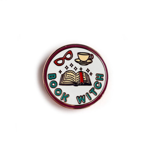 A circular enamel pin in the style of a merit badge featuring illustrations of an open book with sparkles, a teacup, and red glasses. Underneath the illustrations are the words "Book Witch" in teal bubble letters.